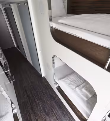 Moderne interiør i et kapselhotell, med elegante, hvite rom med pent oppredde senger stablet i køyestil langs en gang med tregulv. Frostete glassdører er synlige i enden av korridoren.