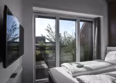 Et moderne soverom med en pent oppredd dobbeltseng. En flatskjerm-TV er montert på veggen. Store glassdører åpner ut til en balkong med grøntområder, under en klar blå himmel. Naturlig lys lyser opp rommet.