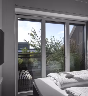 Et moderne soverom med en pent oppredd dobbeltseng. En flatskjerm-TV er montert på veggen. Store glassdører åpner ut til en balkong med grøntområder, under en klar blå himmel. Naturlig lys lyser opp rommet.