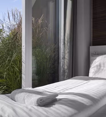 En pent oppredd seng med hvitt sengetøy og et sammenbrettet håndkle er plassert ved en stor skyvedør i glass. Sollys kaster skygger på sengen. Utenfor er frodige grønne planter og en klar blå himmel synlig gjennom glasset.
