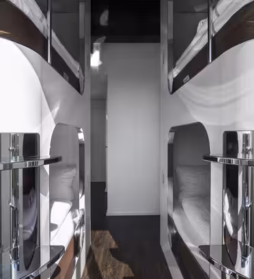 Et moderne, elegant vandrerhjem med to rader med hvite soveputer. Hver pod har en kromfinish, sengetøy og et vindu. Gulvet er mørkt tre, og den generelle atmosfæren er ren og minimalistisk.