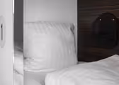 Et nærbilde av en koselig seng med hvite puter og laken inne på et kapselhotell. En liten svart pose er festet til trepanelet ved siden av sengen, og et nummer "1" er synlig på kapseldøren.