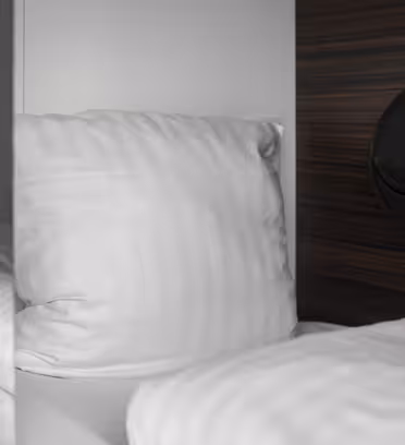 Et nærbilde av en koselig seng med hvite puter og laken inne på et kapselhotell. En liten svart pose er festet til trepanelet ved siden av sengen, og et nummer "1" er synlig på kapseldøren.