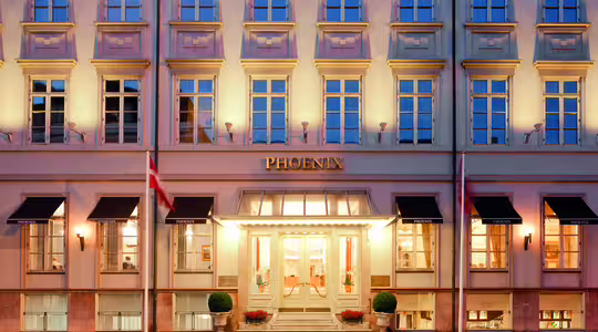 Bildet viser det elegante eksteriøret til Phoenix Hotel, med en klassisk fasade med flere rader med opplyste vinduer. Inngangen er storslått, med to flagg som flankerer døråpningen og potteplanter ved trappen.