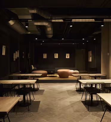 Et svakt opplyst moderne interiør med trebord og svarte stålstoler. Rommet har en minimalistisk design, med kunst på veggene og en koselig sittegruppe med rosa polstrede stoler i bakgrunnen. Utsatte industrielle himlingselementer er synlige.