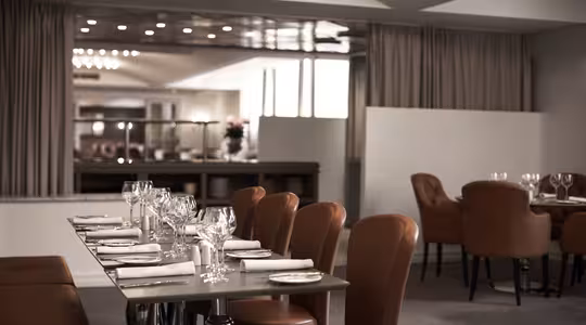 Et elegant restaurantinteriør med pent dekkede bord, med brune skinnstoler og hvite servietter. Myk belysning skaper en varm atmosfære. Bakgrunnen viser store gardiner og speil, noe som forbedrer plassen.