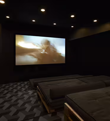 En svakt opplyst hjemmekino med en stor skjerm som viser en uskarp actionscene. Rommet har flere polstrede sittegrupper på mønstrede gulv, omgitt av mørke vegger med subtil belysning over hodet.