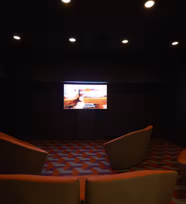 Et svakt opplyst rom med fire oransje stoler vendt mot en stor skjerm som viser en film eller presentasjon. Gulvet har et geometrisk rødt og grått mønster. Taklamper lyser opp rommet mykt.