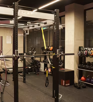 Et treningsstudio med knebøystativ, vektstang, treningssykkel, kettlebells i ulike farger, medisinballer og vektskiver. Plassen er godt opplyst, med speil og svart gulv. Hengende stropper er festet til stativet for ekstra øvelser.
