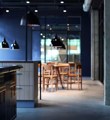 Moderne kjøkken og spisestue i industriell stil med synlige betongvegger og -tak. Svarte pendellamper henger over et spisebord i tre med stoler, og en elegant benkeplate med metalliske detaljer er i forgrunnen. Naturlig lys filtrerer inn.