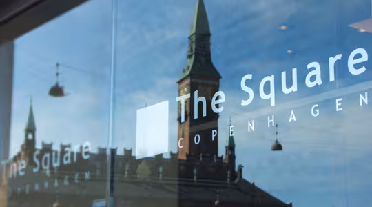 Refleksjon av en historisk bygning med et tårn i glassvinduet til «The Square Copenhagen», med klarblå himmel og gatelys.