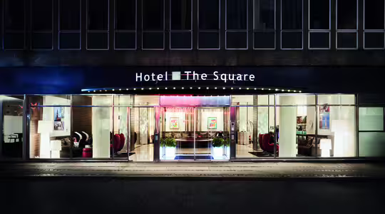 Bildet viser den opplyste inngangen til Hotel The Square om natten. Fasaden er i glass med synlige sittegrupper innvendig. Hotellnavnet vises tydelig over inngangen.