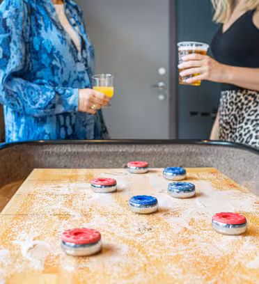 To kvinner står ved et shuffleboard-bord og holder drinker. Bordet er drysset med hvitt pulver og har røde og blå pucklignende brikker. Begge kvinnene er uformelt kledd, og settingen fremstår avslappet og sosial.