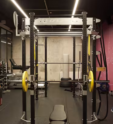 Et treningsstudio med strømstativ utstyrt med vektstang og gule vektskiver. Det er en benk under stativet, kettlebells og vekter på stativer til venstre, en rosa vegg med strekillustrasjoner og diverse treningsutstyr rundt.
