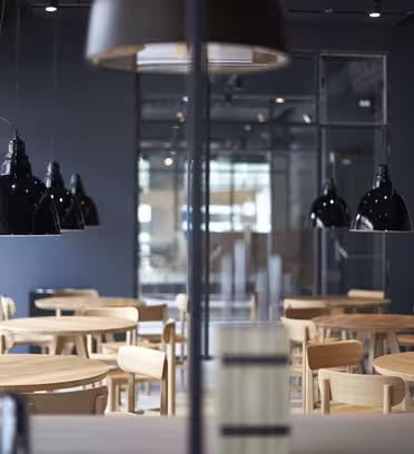 Et moderne kaféinteriør med runde trebord og matchende stoler. Svarte pendellamper henger i taket og kaster lys på bordene. Veggene er mørke, og store vinduer for naturlig lys er synlige i bakgrunnen.