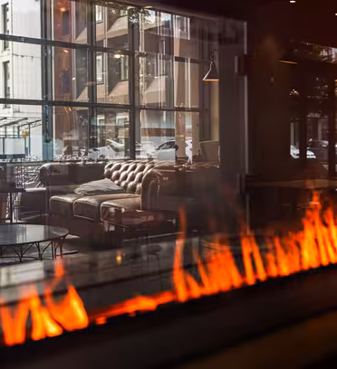 Refleksjoner av et koselig salongområde med skinnsofaer og moderne bord er synlige gjennom et glasspanel. En flammeeffekt er tilstede på glasset, noe som antyder en varm og innbydende atmosfære innendørs.