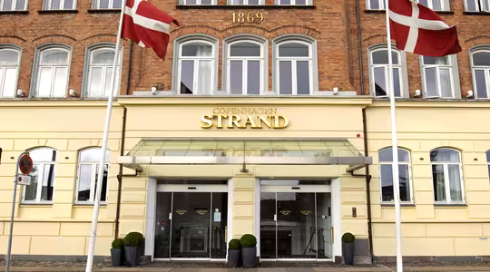 Bildet viser inngangen til Copenhagen Strand Hotel, en murbygning med store vinduer og årstallet 1869 vist ovenfor. To danske flagg er plassert på hver side av inngangen.