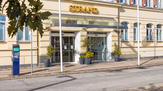 Inngang til Copenhagen Strand hotell med glasstak, store vinduer og potteplanter. Parkeringsautomat og flaggstenger med røde flagg er synlig på fortauet. En bladrik gren er i forgrunnen.