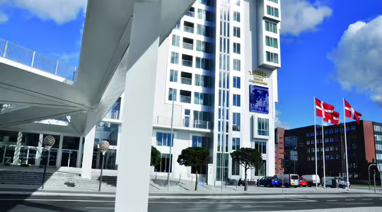 Et moderne hvitt høyhus med unik design står under en hvit overgang. Tre danske flagg vaier i nærheten. Biler står parkert langs gaten, og en klarblå himmel med spredte skyer danner bakteppet.