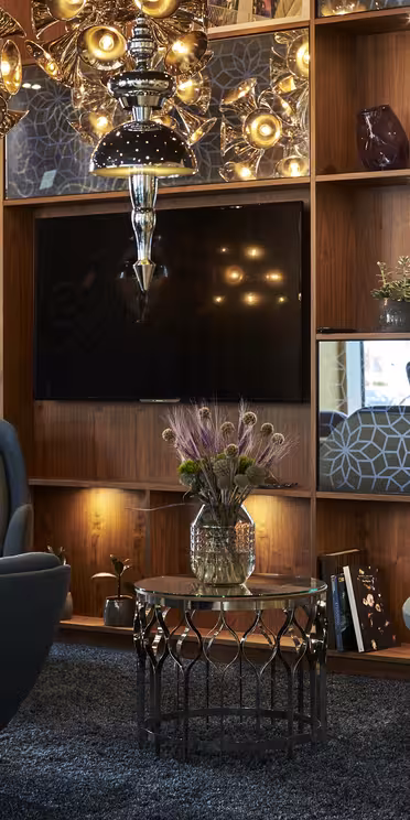 En koselig stue med en bokhylle i tre med geometriske design, planter og ulike dekorative gjenstander. To blå lenestoler og et rundt salongbord i glass er sentrert under en moderne lysekrone. En TV er montert i hyllene.