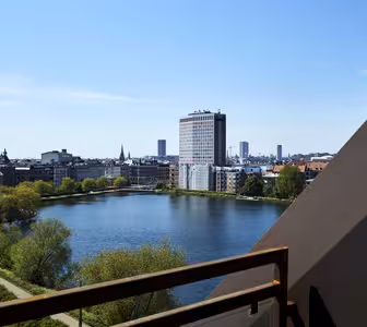 En utsikt over et bybilde med en rolig innsjø, omgitt av bygninger. En høy, moderne bygning med et fossefall står fremtredende. Bakgrunnen viser en klar, blå himmel og en fjern byhorisont.