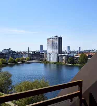 En utsikt over et bybilde med en rolig innsjø, omgitt av bygninger. En høy, moderne bygning med et fossefall står fremtredende. Bakgrunnen viser en klar, blå himmel og en fjern byhorisont.