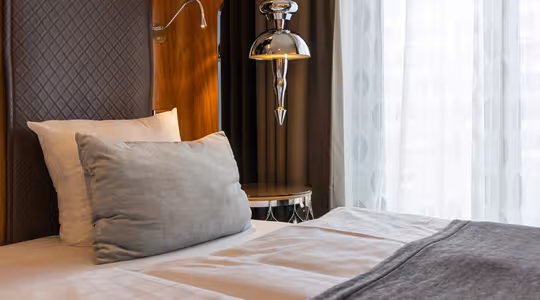 Et koselig hotellrom med en pent oppredd seng med hvitt og grått sengetøy, en stor grå pute og en moderne sølvlampe. Sengen er plassert ved siden av et vindu med rene hvite gardiner, slik at naturlig lys slipper inn i rommet.