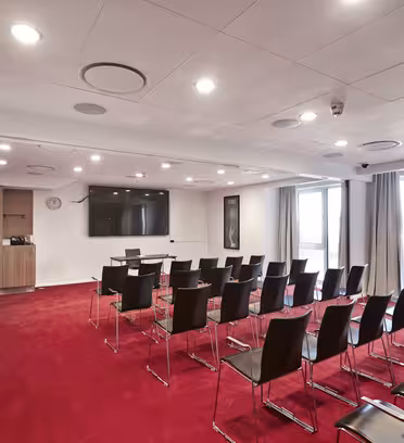 Et konferanserom med rød løper, arrangert med rader med svarte stoler vendt mot en stor TV-skjerm montert på veggen. Et podium og et lite bord med stoler står foran. Rommet har beige vegger, taklys og store vinduer med gardiner.