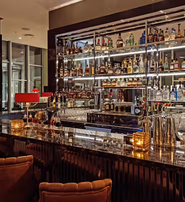 En stilig bar med benkeplate i marmor og røde lamper. Hyller bak baren viser frem ulike brennevinsflasker. Komfortable brune barkrakker langs disken. Varm belysning og dekorative elementer skaper en innbydende og elegant atmosfære.