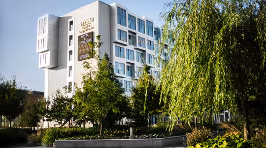 Moderne hvit bygning omgitt av frodige grøntområder og trær, med store vinduer. Et skilt med "Hotel Evolution" er synlig på fasaden. Forgrunnen inkluderer en steinsti og anlagt hage. Himmelen er klar og blå.