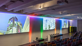 Et moderne konferanserom med fargerike lyseffekter langs veggene. Seks stoler er arrangert foran en stor skjerm som viser arkitektoniske bilder. Fargesterke seter er arrangert i rader mot skjermen.